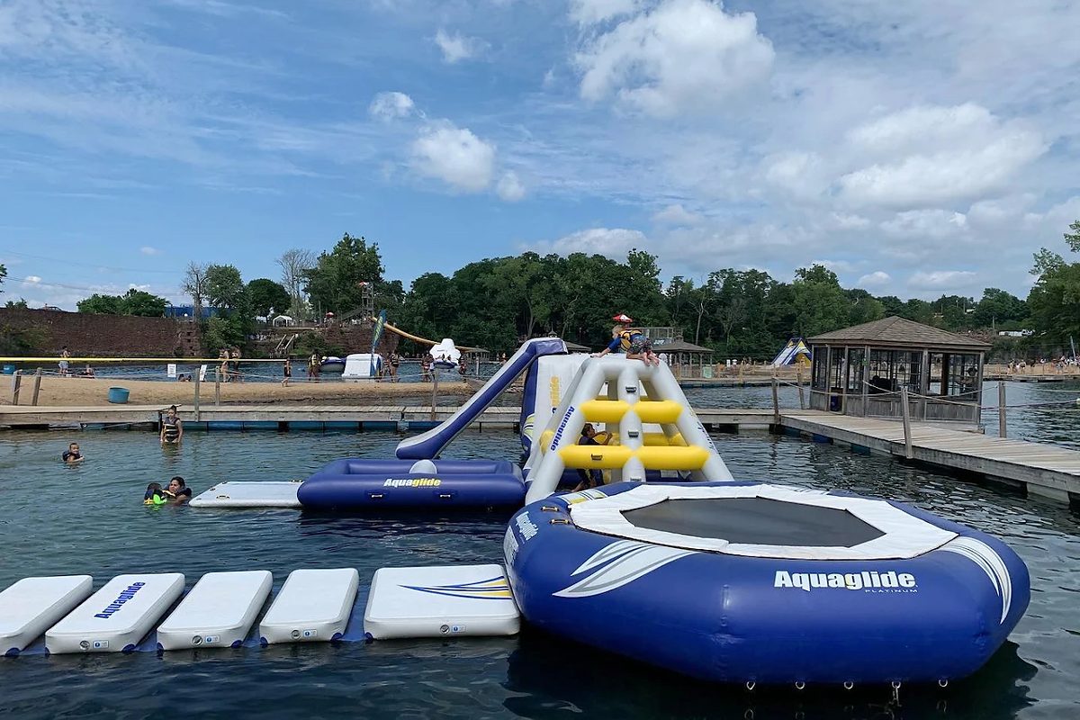 Swimmers and inflatable floating obstacles on the green water of a flooded brownstone quarry, with a sandy beach and the cut quarry wall behind
