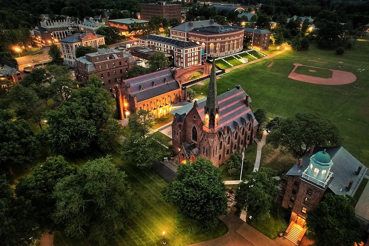 Wesleyan University from the air at dusk, the lit brownstone chapel and its spire in the foreground with playing fields and campus buildings beyond