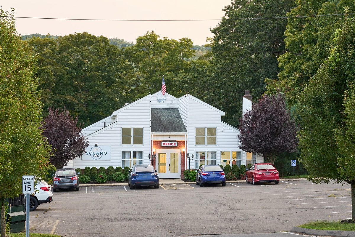 The Solano Inn front office at dusk: a white clapboard building with a lit OFFICE sign over the entrance, the Solano Inn sign on the wall beside it, and guest cars parked directly outside