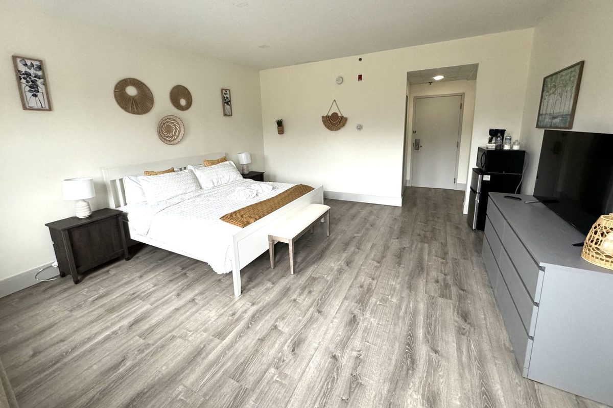 A king guest room with a white platform bed, dark wood nightstand, woven basket wall decor and a low dresser holding a flat-screen TV beside the entry door.