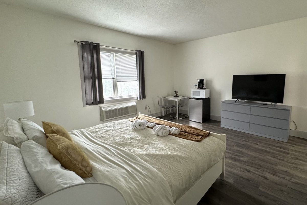 A king room with a coffee maker, microwave and compact refrigerator arranged on a desk beside the window, and a flat-screen TV on a grey dresser opposite the bed.