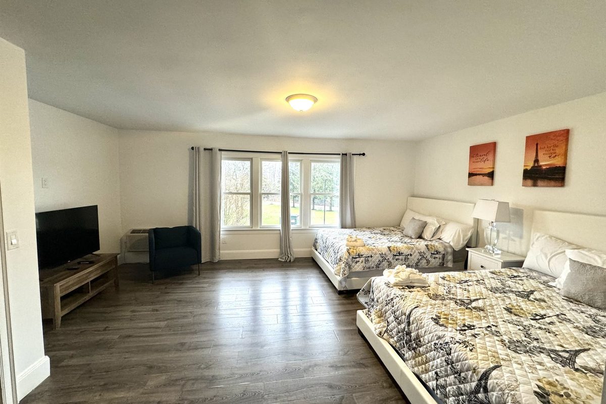 A double queen room with a wall-mounted flat-screen TV, a blue armchair by the window and two queen beds with patterned quilts on a wood-look plank floor.