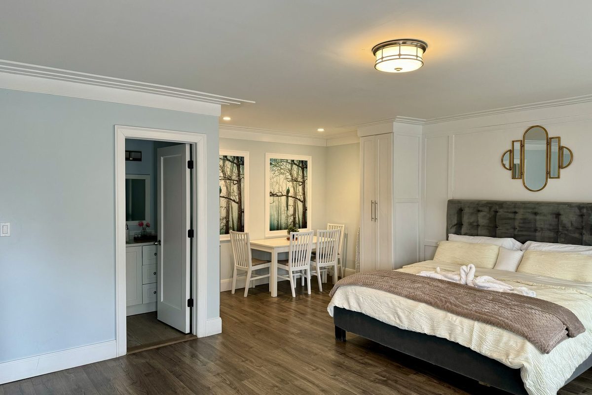 A king room with a grey upholstered headboard, an open doorway through to the bathroom, and a white dining table with four chairs beside the window.