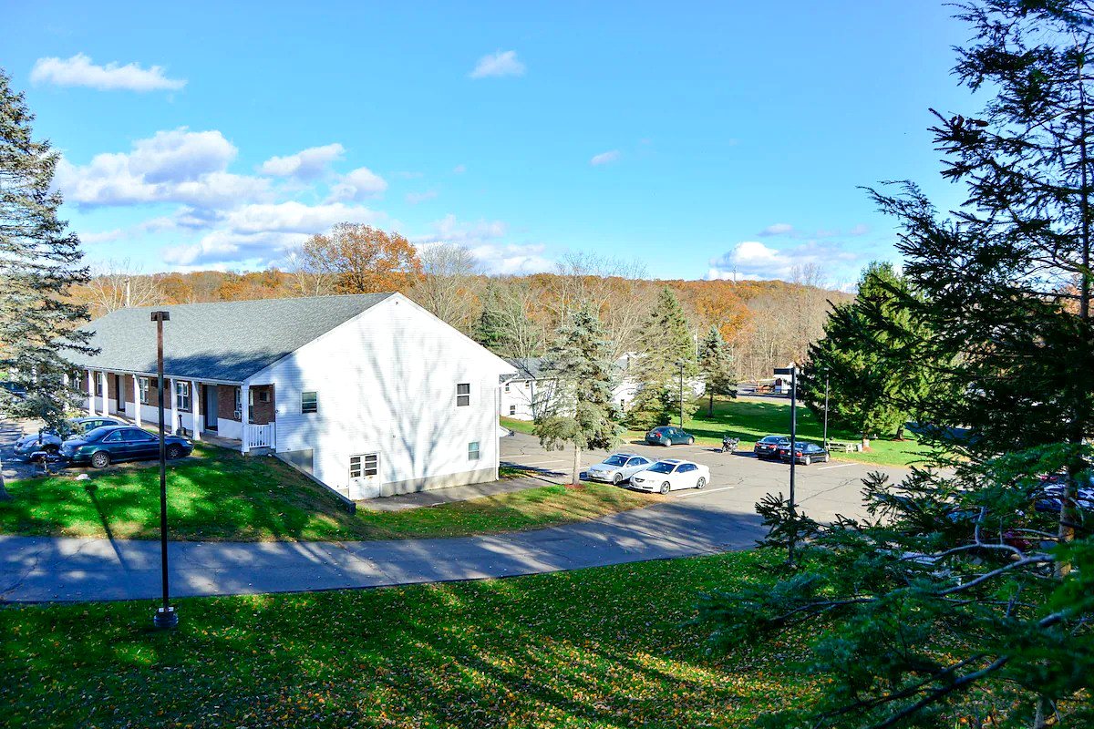An elevated view across the lawn in autumn, with a guest building, parked cars and evergreens in the foreground and a ridge of orange and gold trees on the horizon under a blue sky.
