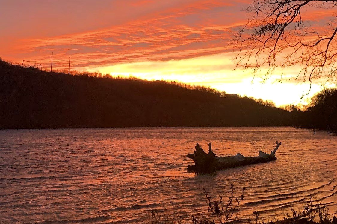 Sunset over the Connecticut River, with an orange and red sky above a dark wooded ridge, bare branches framing the right of the frame and a fallen log in the shallow water. One mile from the property.
