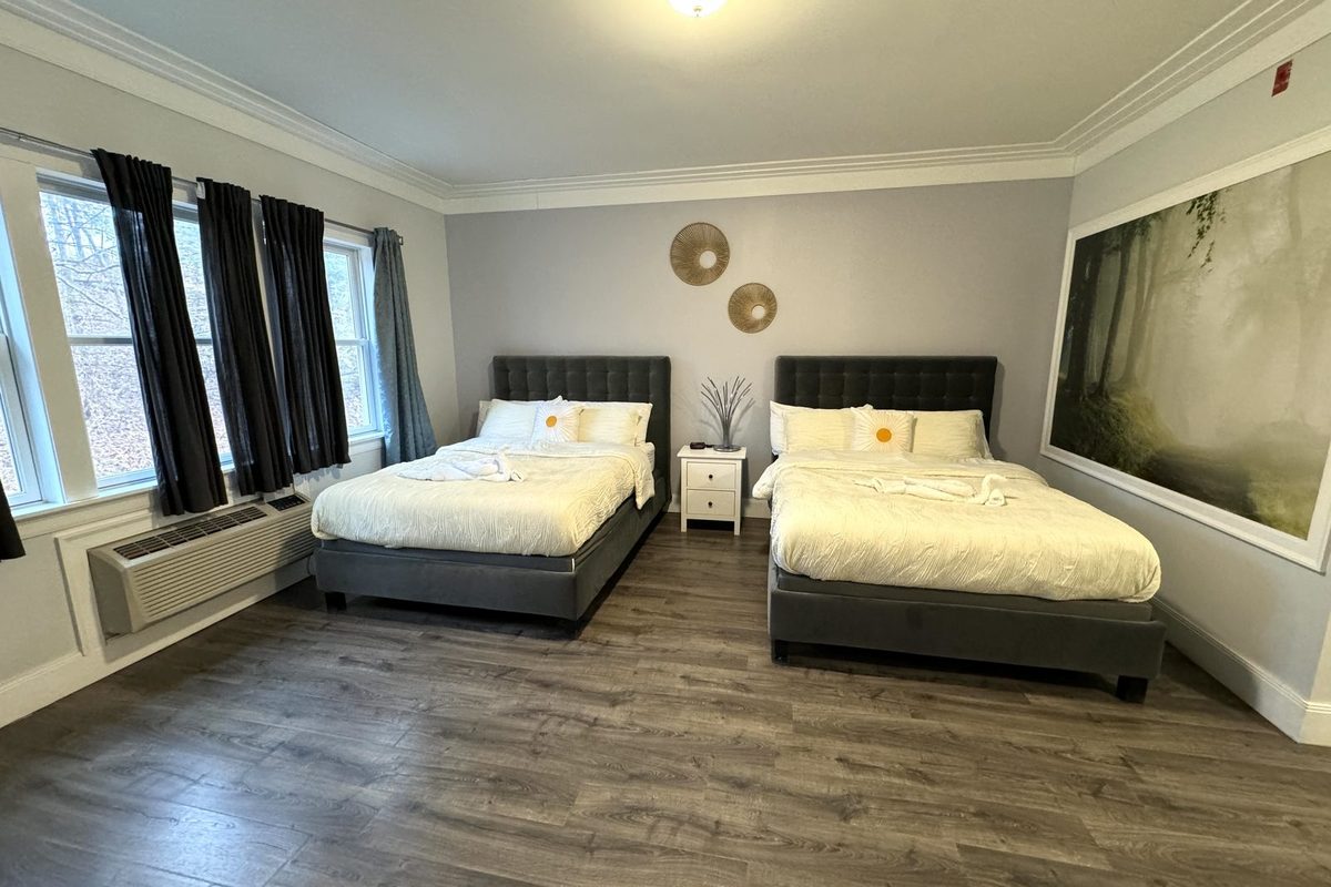 Two queen beds with grey upholstered headboards and a white nightstand between them, blackout curtains at the windows and a through-wall air conditioner below.
