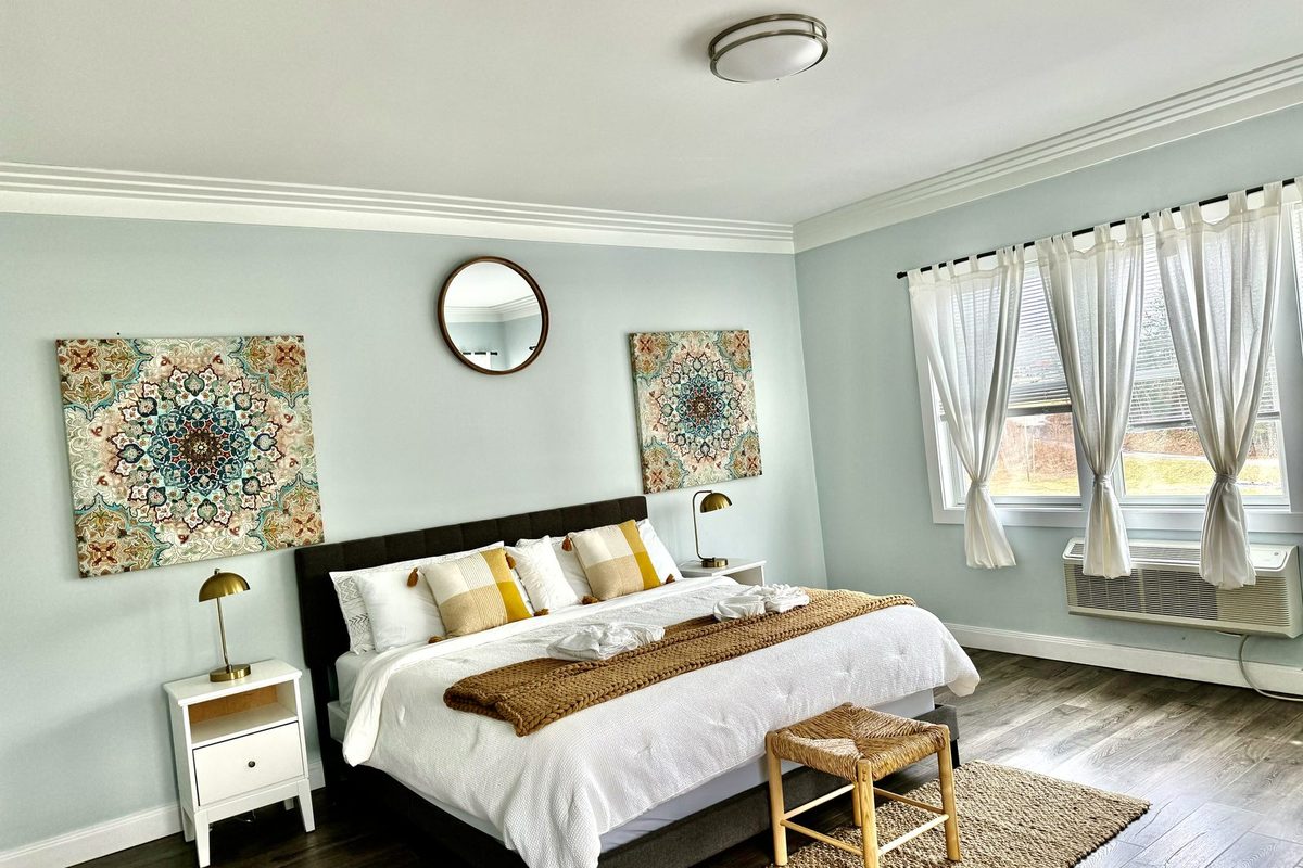 King bed dressed in white and gold linens against a pale blue wall, with framed mandala prints and a round mirror above the headboard, a woven bench at the foot of the bed, and a curtained window with a through-wall air conditioner.